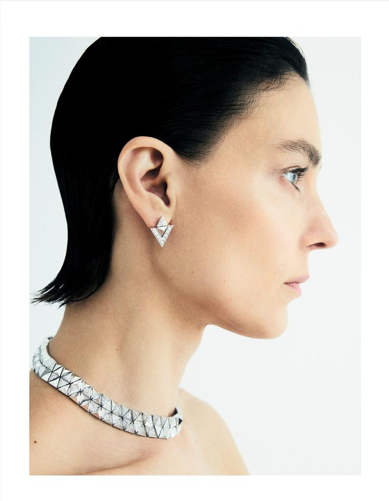 Profile view of an adult female with dark hair wearing geometric diamond necklace and earrings against a plain white background.