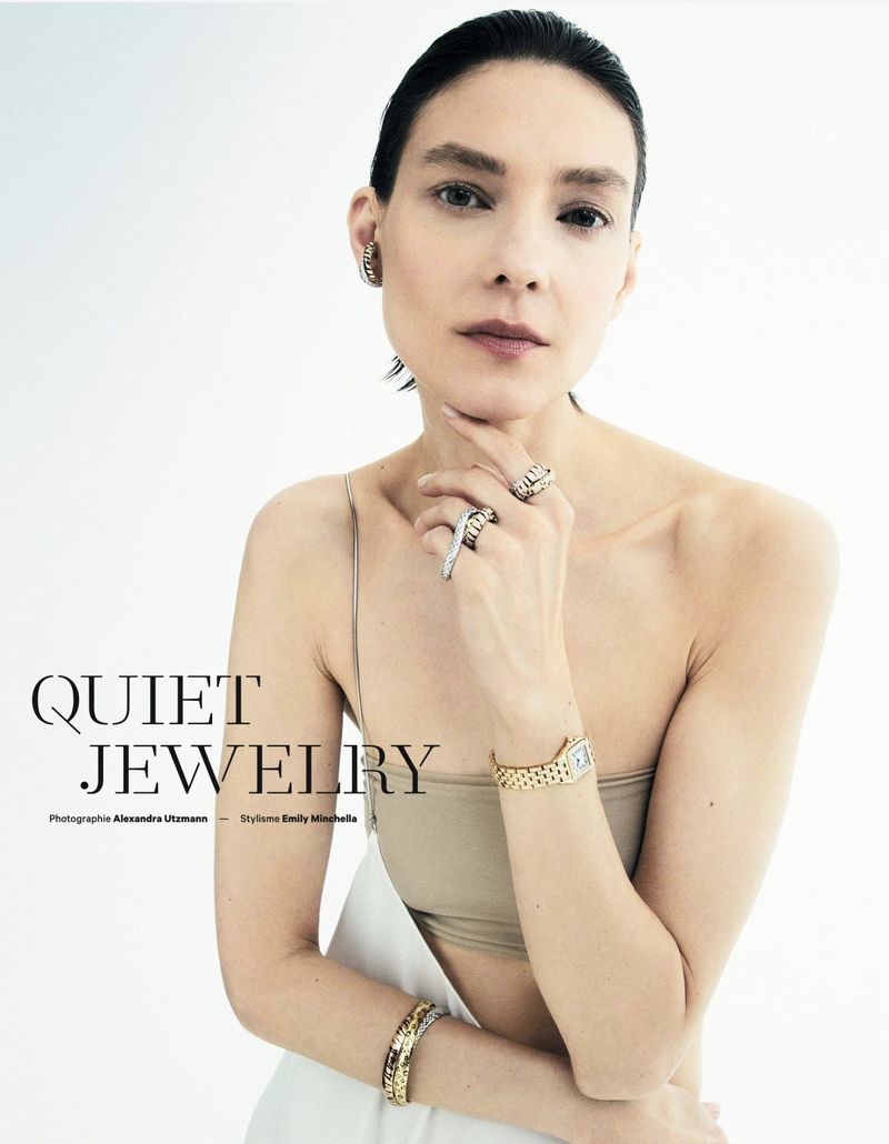A woman in a studio wearing jewelry, including a watch, rings, and a bracelet, with a neutral top and white bottoms, and soft lighting.