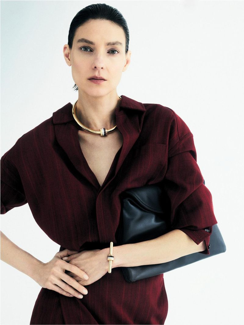Brunette woman in maroon shirt dress holds black leather clutch purse and wears gold choker and bracelet against white backdrop.