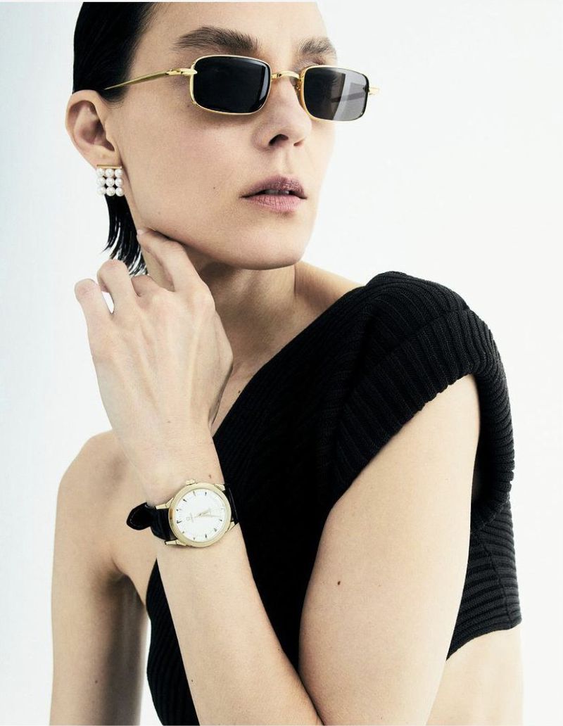 A young adult female model wears rectangular sunglasses, pearl earrings, and a black top, posing in a studio with a wristwatch.