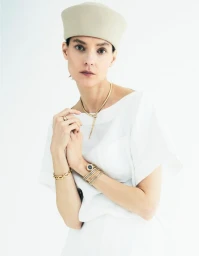 Model wearing a neutral beret, white top, and gold jewellery against a white studio background.
