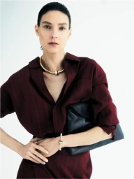 Model Kati Nescher wearing a burgundy dress and gold jewellery, holding a black handbag against a white background