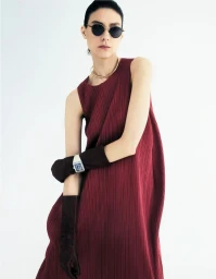 Model Kati Nescher wearing a pleated burgundy dress, sunglasses, and long brown gloves against a white studio background.
