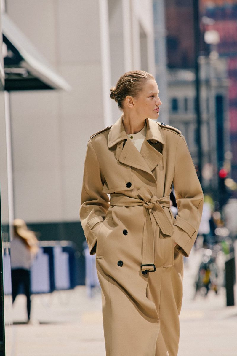 Adult woman in a beige trench coat walking in an urban setting with blurred buildings in the background, captured in an eye-level shot.