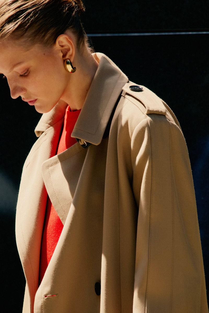 Woman poses in a tan trench coat with brass earrings and red shirt against a dark background in a close-up shot.