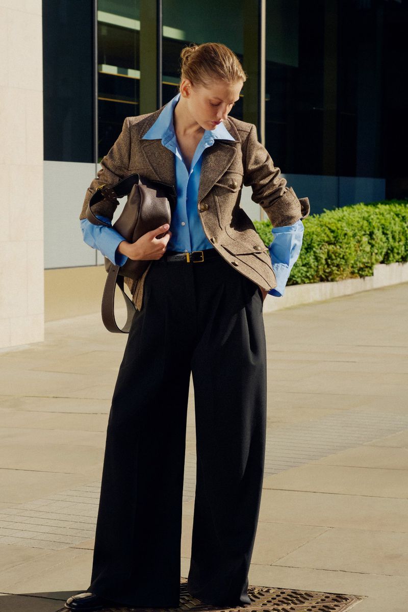A woman in a blazer and wide-leg pants stands outdoors, carrying a bag over her shoulder, with a modern building in the background.