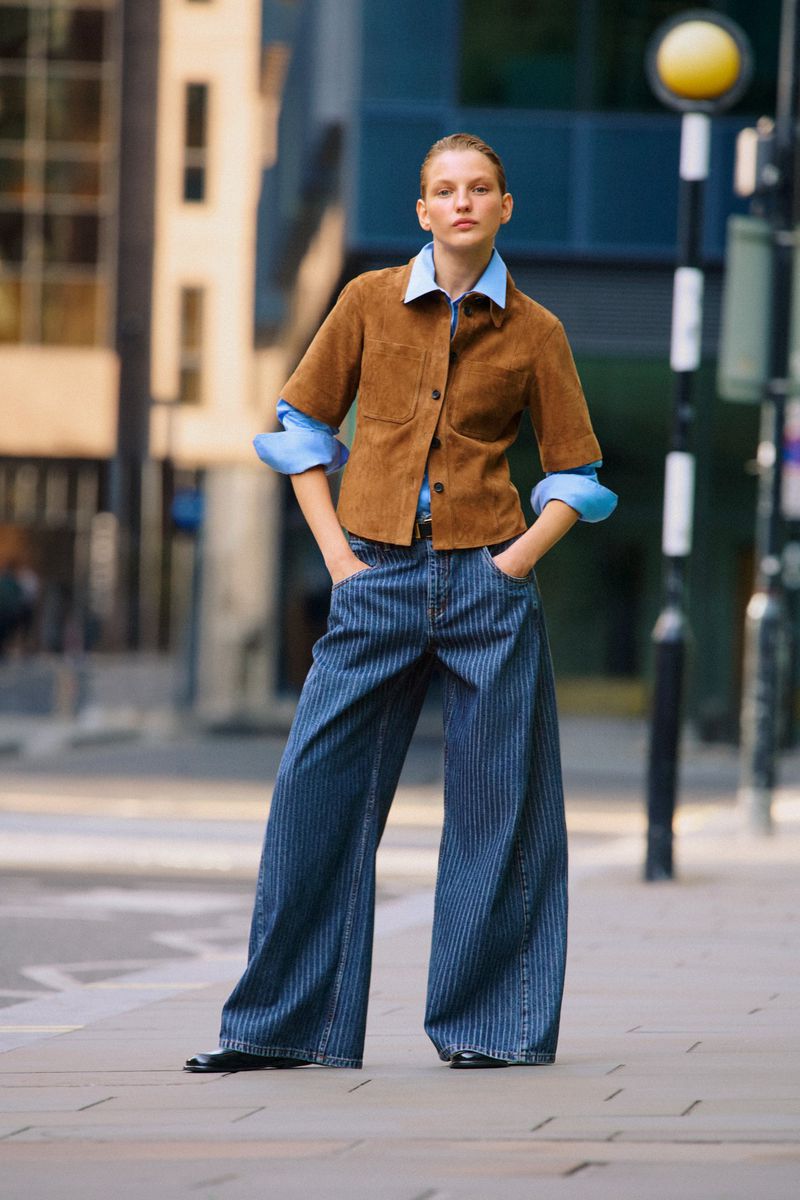 Blonde woman poses in blue striped wide-leg jeans, brown jacket, and blue shirt on an urban sidewalk with a blurred background.