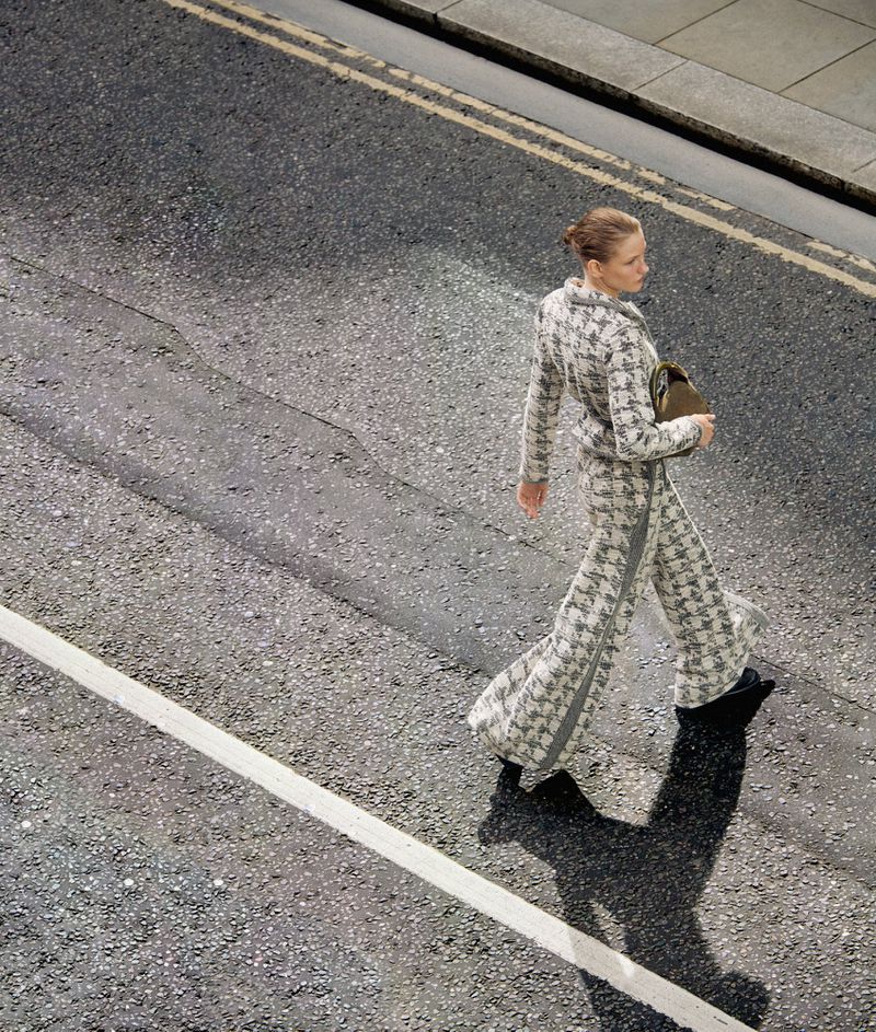 Adult woman in patterned pantsuit crosses street. Top-down, eye-level angle on asphalt road with painted lines, natural light, and shadows.