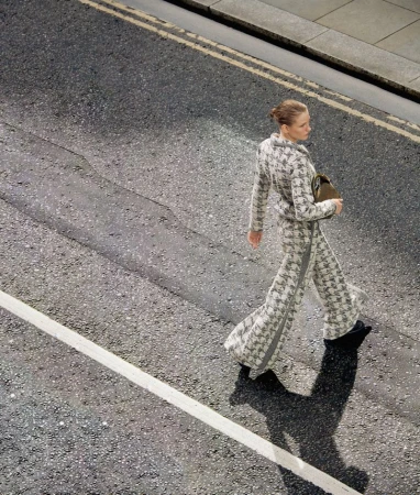 Adult woman in patterned pantsuit crosses street. Top-down, eye-level angle on asphalt road with painted lines, natural light, and shadows.