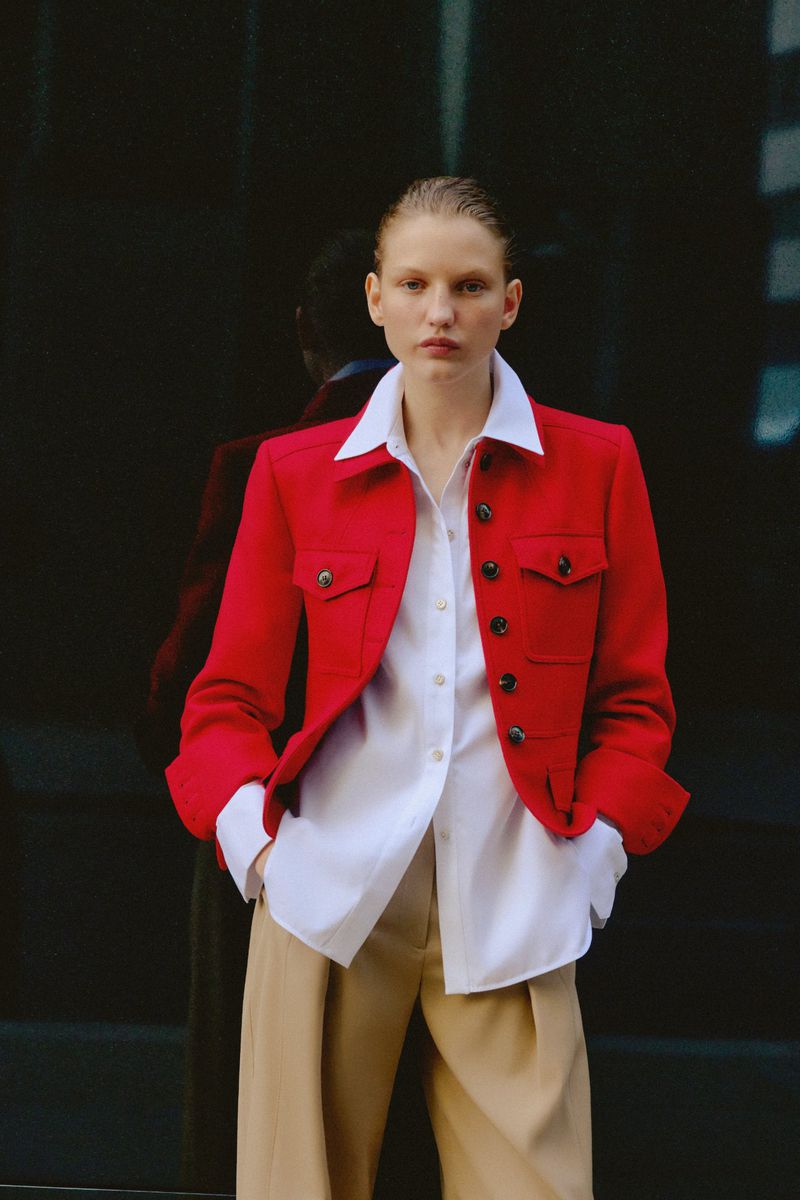 Portrait of an adult female fashion model in a red jacket, white shirt, and tan trousers, posing against a dark reflective background.