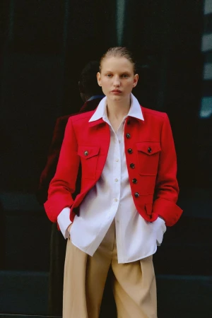 Portrait of an adult female fashion model in a red jacket, white shirt, and tan trousers, posing against a dark reflective background.