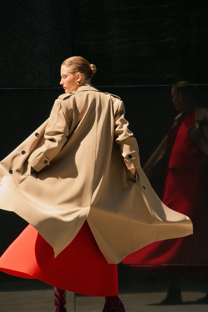 A woman in a tan trench coat and red dress is walking and looking back, casting a reflection on a dark surface in the background.
