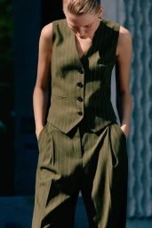 Model wearing an olive pinstripe vest and matching trousers with hands in pockets against a textured background