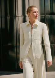 Model wearing a textured cream jacket and matching trousers standing outdoors against a dark panelled glass building.