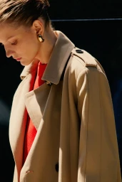 Model wearing a camel trench coat and red top with gold earrings against an outdoor wall