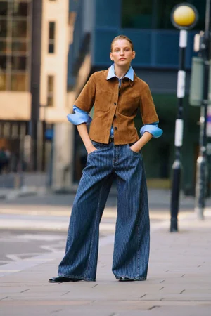 Model standing on a city street wearing a brown suede jacket over a blue shirt and striped wide-leg jeans