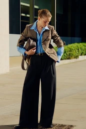 Model wearing a brown tweed jacket over a blue button-down shirt and black trousers holding a dark handbag outdoors.