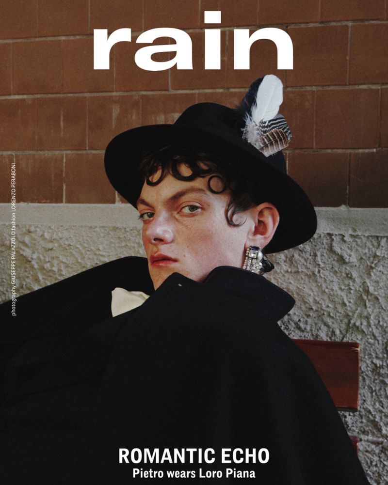 Magazine cover portrait of a young adult with curly hair, wearing a black hat with feather, earring, and black coat in front of a brick wall.