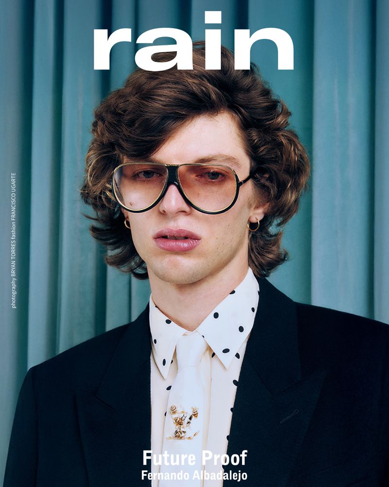 Magazine cover portrait of a young adult male wearing aviator glasses, suit jacket and a polka dot collared shirt, with blue curtains.