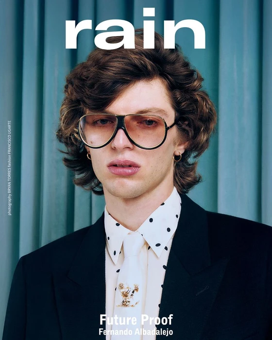 Portrait of a male model wearing tinted aviator sunglasses, a polka-dot collared shirt, and a dark jacket against a vertical ribbed background.