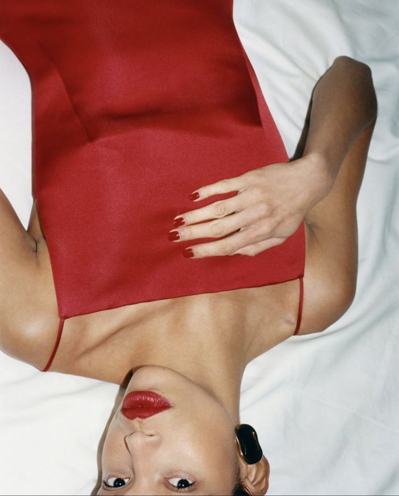 Elegant woman with red lipstick and manicure poses in a red dress, lying on a white sheet for a beauty or fashion shot.