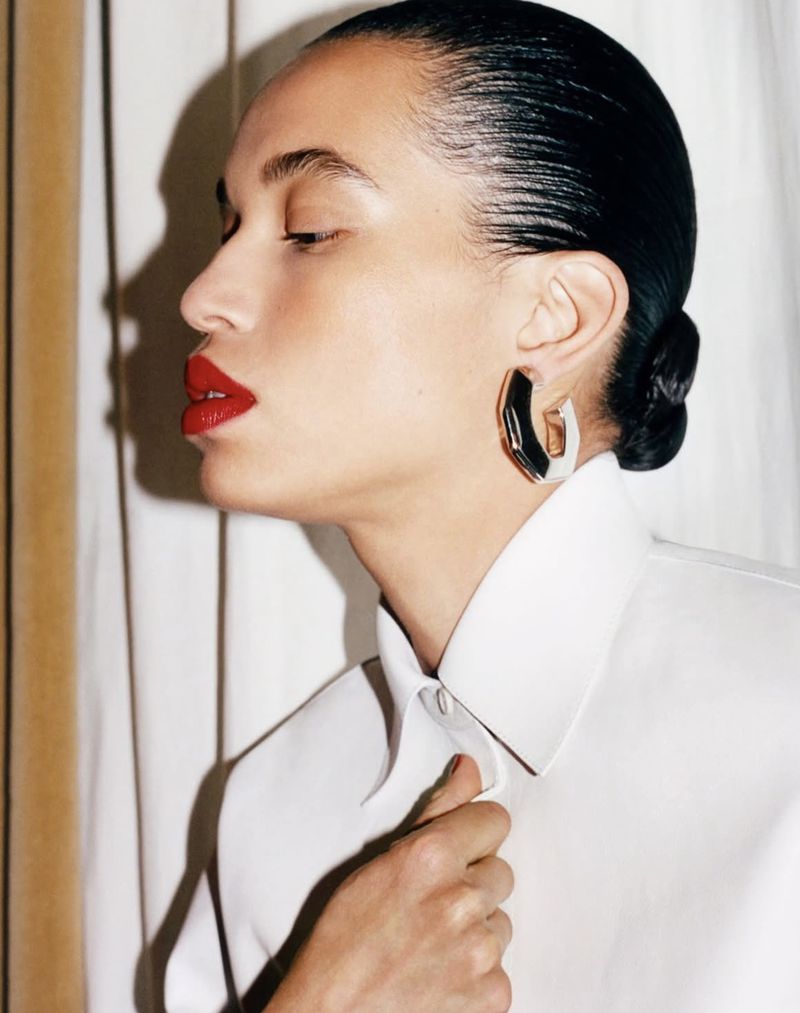 Glamorous headshot of an adult woman with red lips and black hair wearing an elegant white shirt and jewelry in a studio setting.