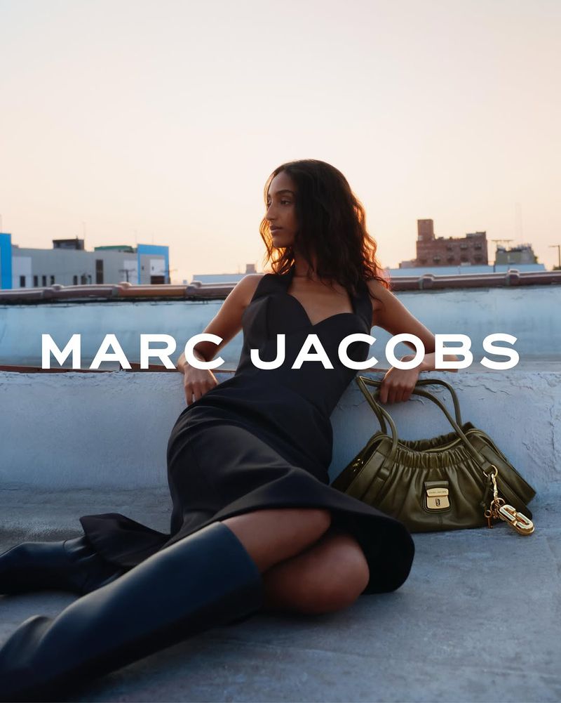 Woman in black dress and boots sitting on a rooftop with a green Marc Jacobs purse, against a cityscape at sunset.