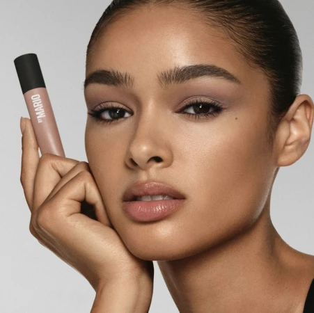 Close-up of a woman with fair skin posing indoors with makeup and a tube of lipstick from By Mario, set against a neutral backdrop.