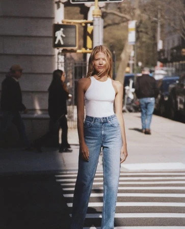 A young woman poses on a crosswalk in the city wearing a white top and jeans, with buildings, trees, and other people in the background.