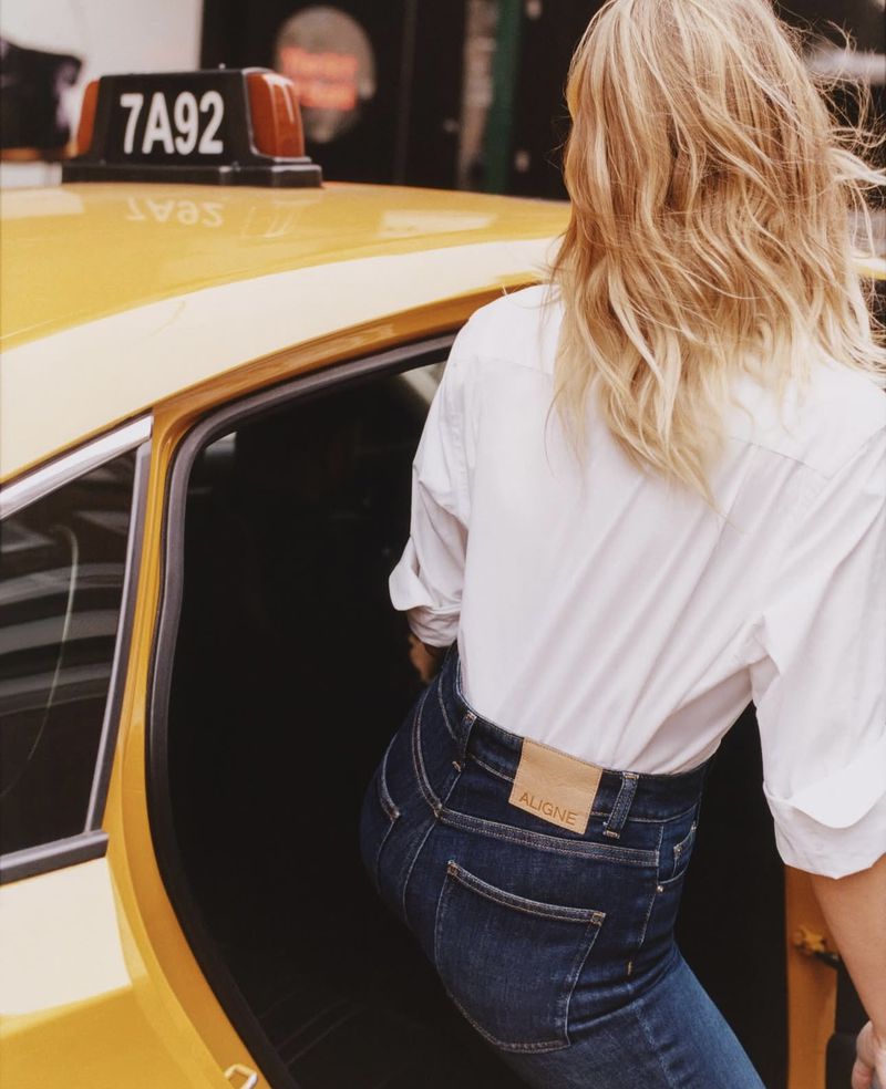 A woman with blonde hair is exiting a yellow taxi, wearing blue jeans and a white shirt, in an urban setting on a bright day.