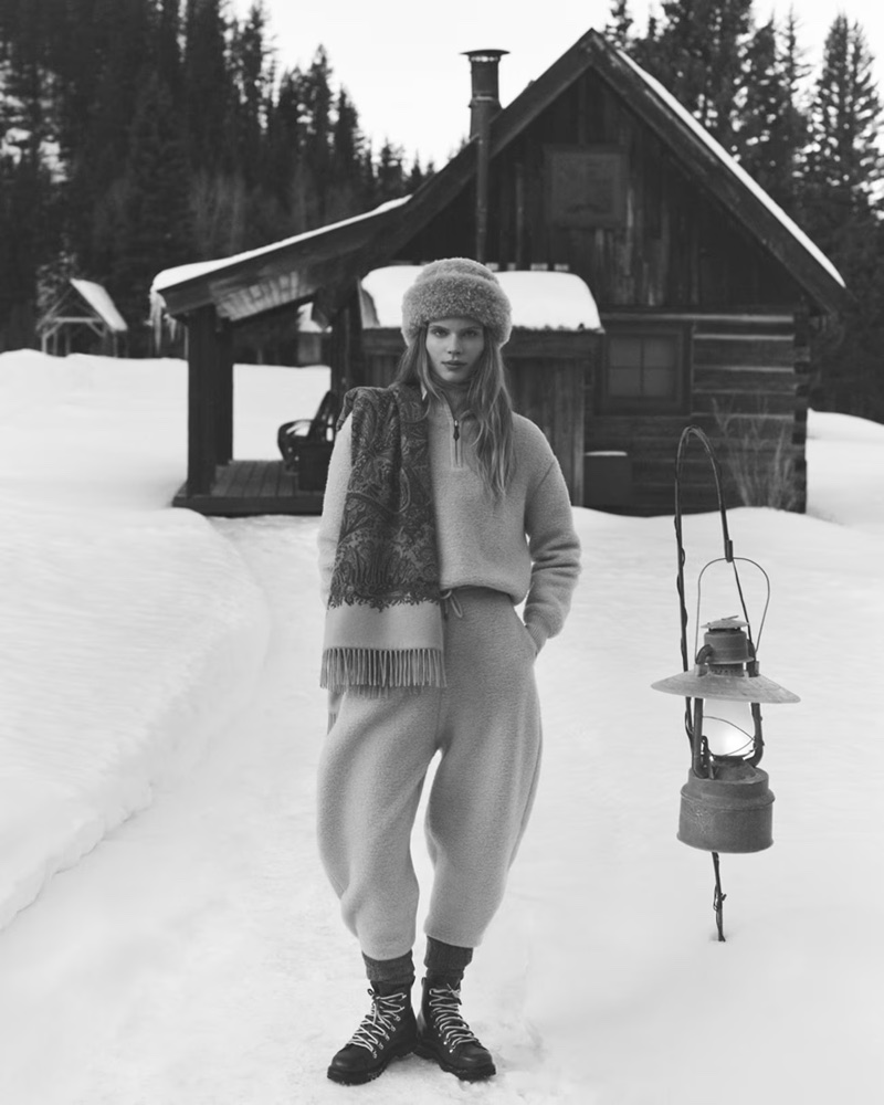Model wearing a fleece pullover, trousers, fur hat, and patterned scarf in front of a snow-covered cabin