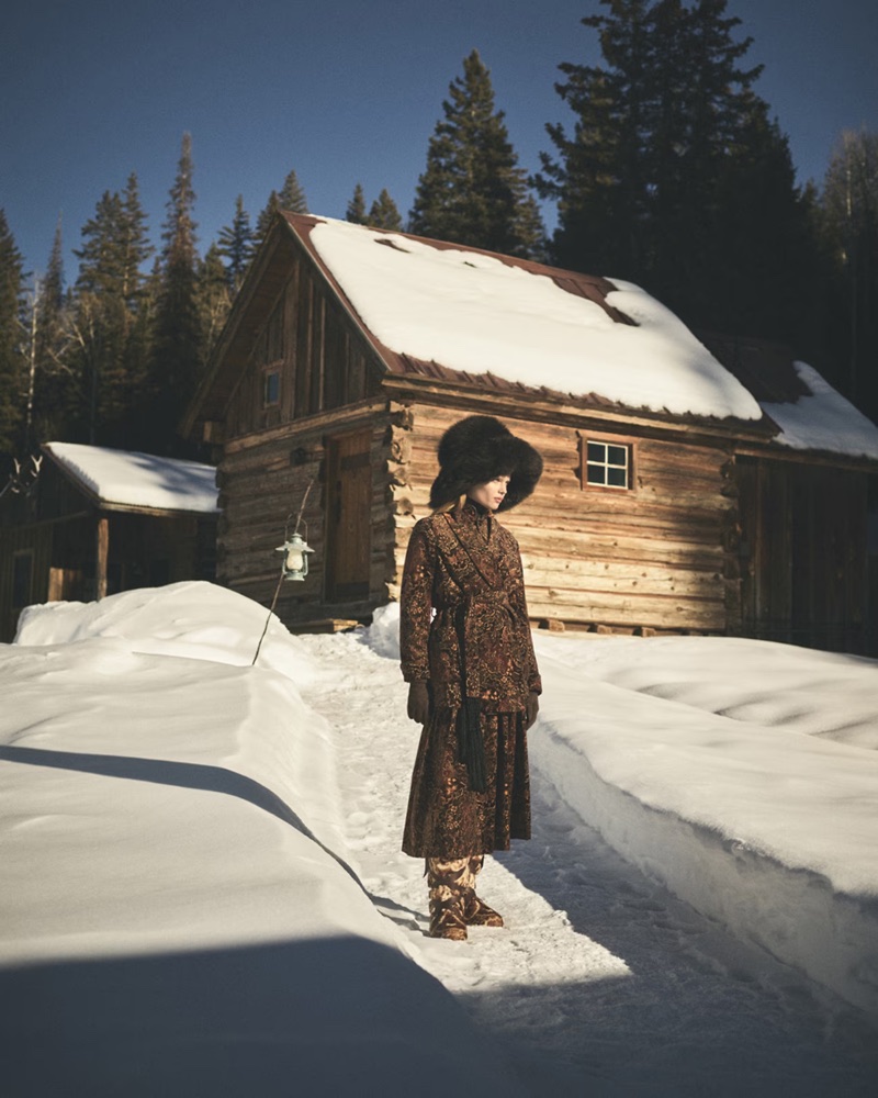 Model wearing a patterned brown coat and fur hat standing outside a snow-covered wooden cabin in a snowy landscape