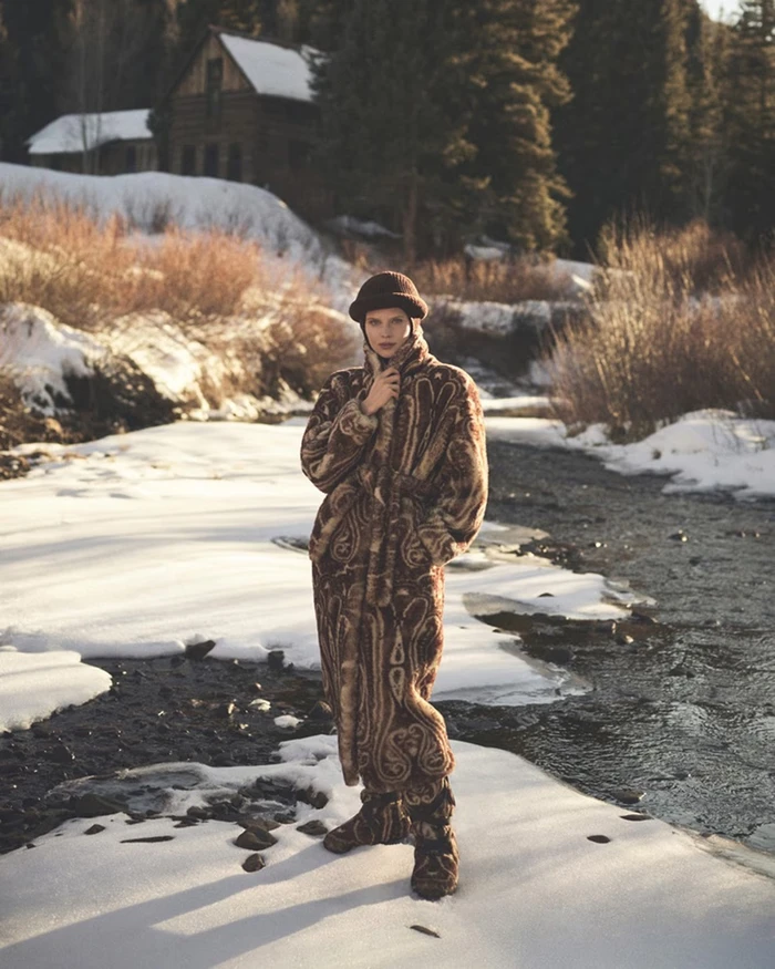 Model wearing a patterned brown fur coat and knitted hat standing on snow beside a frozen river