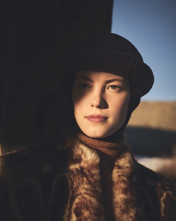 Close-up portrait of a woman wearing a dark knitted cap and a textured fur coat under warm directional sunlight.