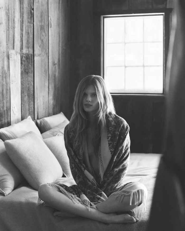 Model seated cross-legged on a bed draped in a patterned fringed shawl with a rustic wood paneled wall and window behind