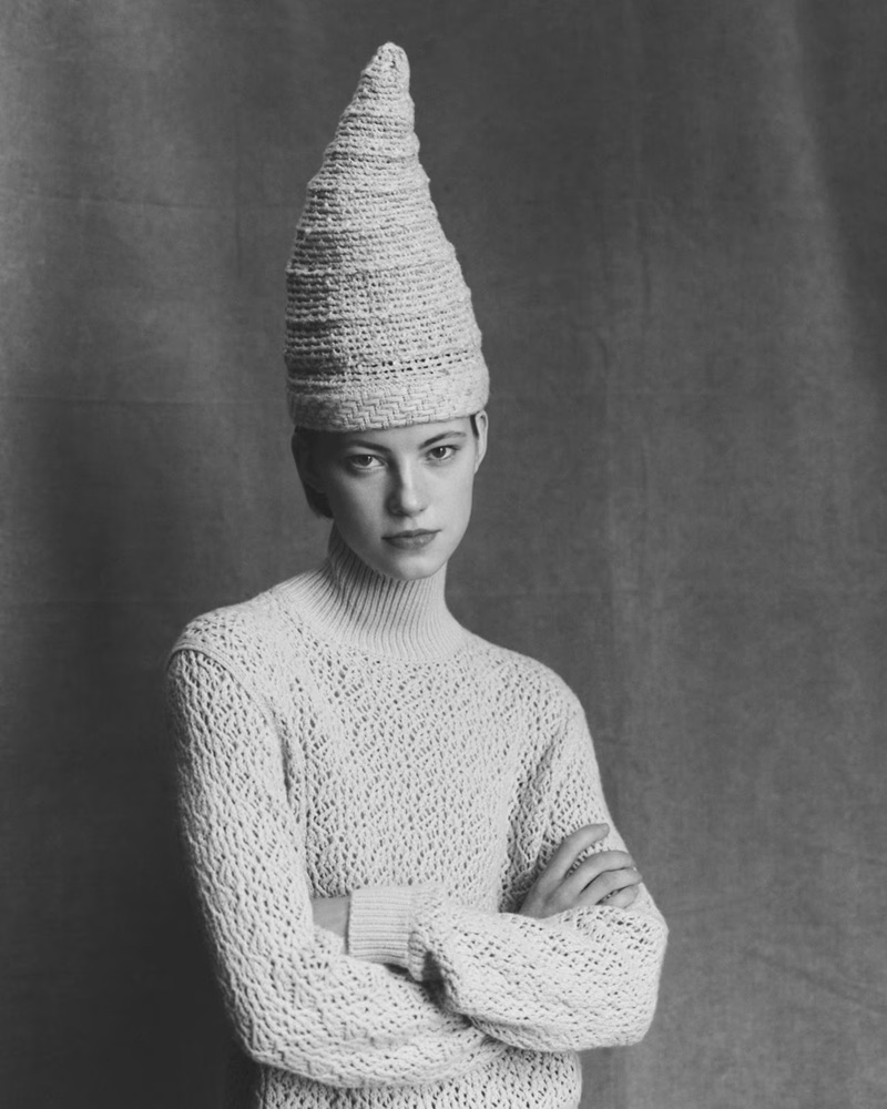 Monochrome portrait of a model wearing a tall textured conical hat and a patterned knit sweater with arms crossed against a textured backdrop