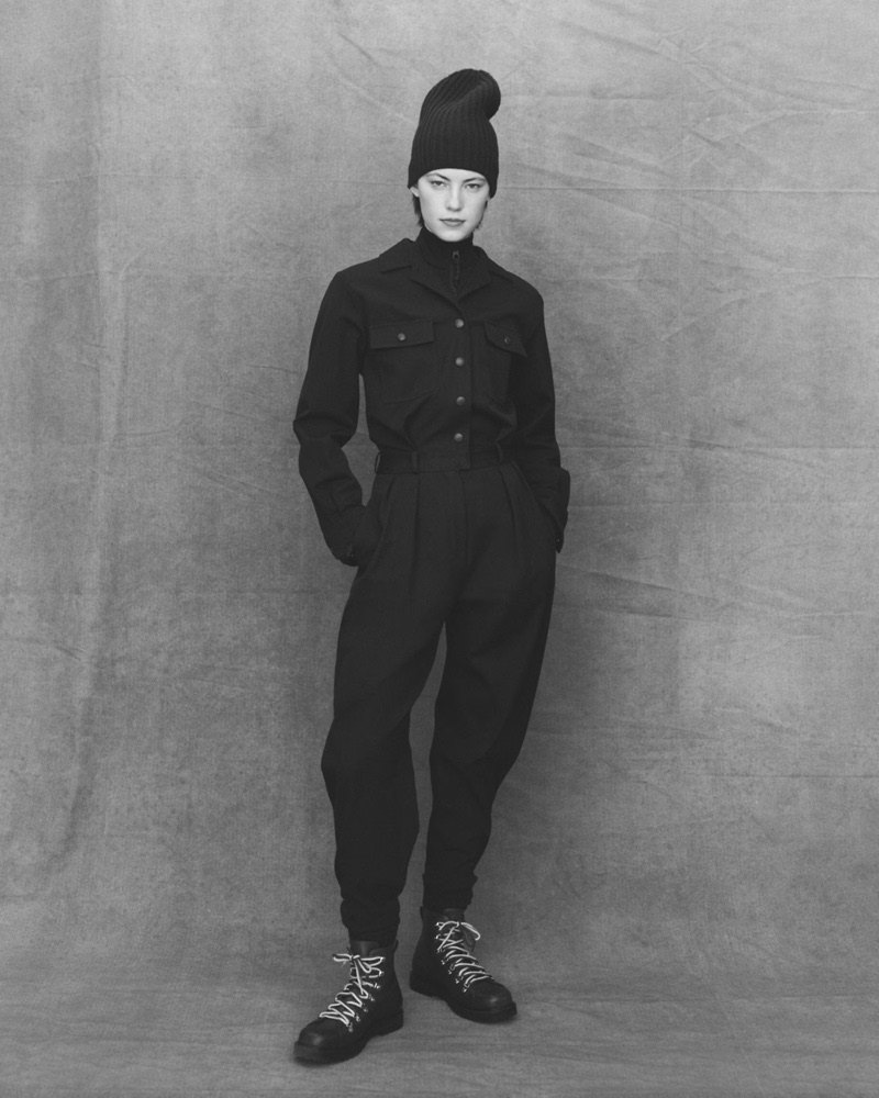 Model in black utility jumpsuit and tall beanie standing against textured backdrop