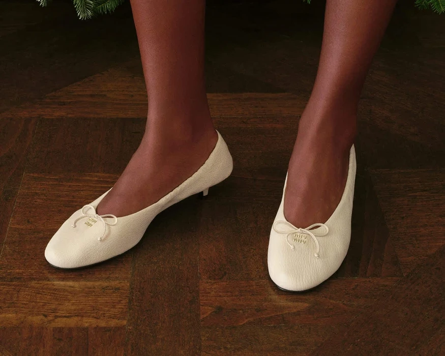 Close-up of a person wearing cream low-heeled ballet flats with small bows on dark parquet flooring for Miu Miu