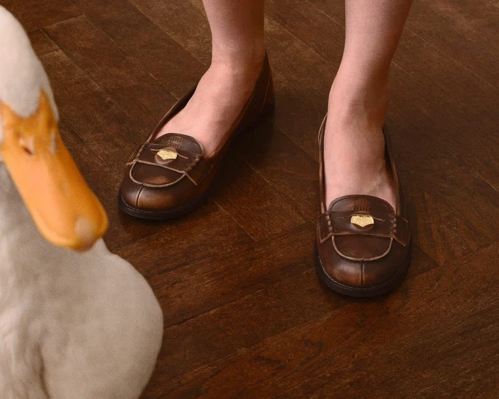 Close-up of a person wearing brown leather loafers on a dark wood parquet floor with a white stuffed animal in the foreground