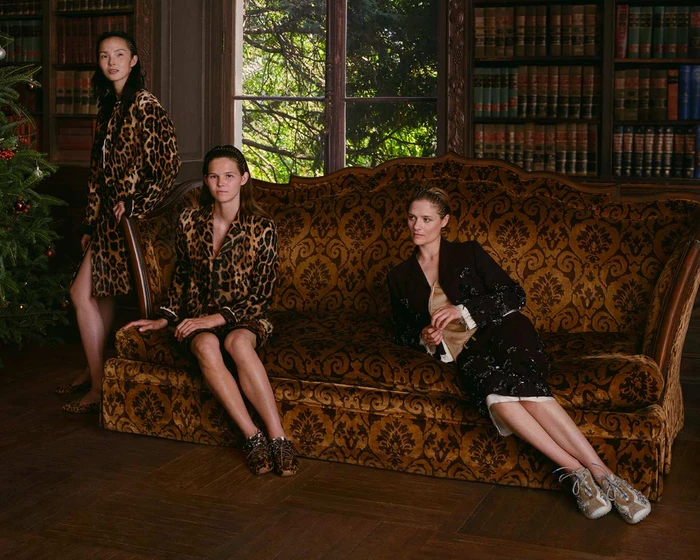 Three female models posing on a large patterned velvet sofa inside a wood-paneled library with floor-to-ceiling bookshelves.