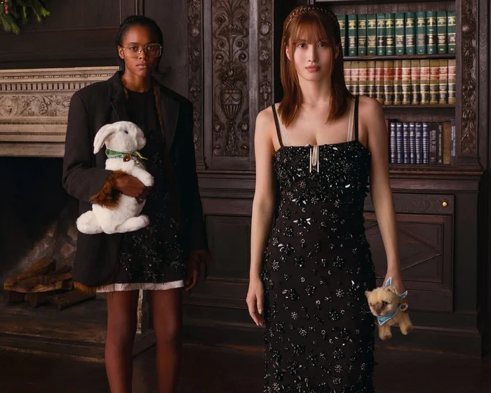 Two models posing in a dark library interior holding plush animal toys for Miu Miu Holiday 2026 campaign.