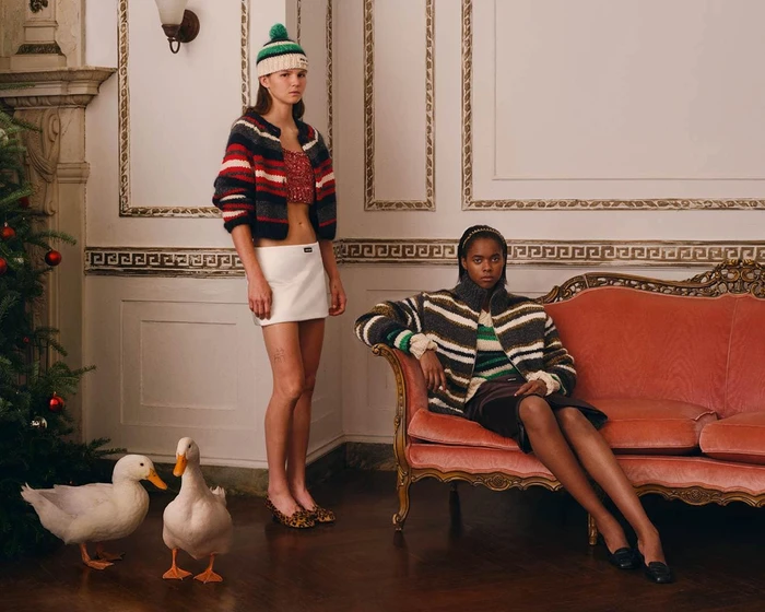 Two models wearing striped knitwear in an ornate room with a red velvet sofa and two white ducks on the floor