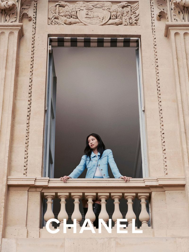 Fashion shot of an adult female, looking out of a stone window in a light blue jacket with Chanel logo at the bottom of the frame.