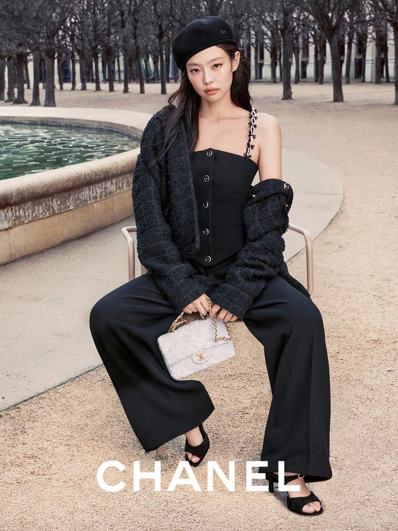 A woman wearing a black beret and a two-piece black outfit poses with a purse in an outdoor setting, with the brand CHANEL visible.