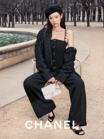 A woman wearing a black beret and a two-piece black outfit poses with a purse in an outdoor setting, with the brand CHANEL visible.