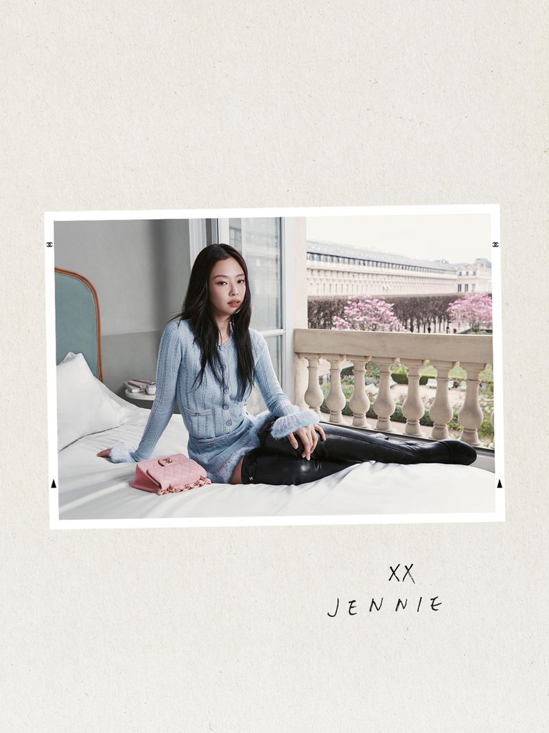 A woman poses on a bed in front of a window, showcasing a light blue knit set with glossy black boots and a pink purse. Balcony visible.