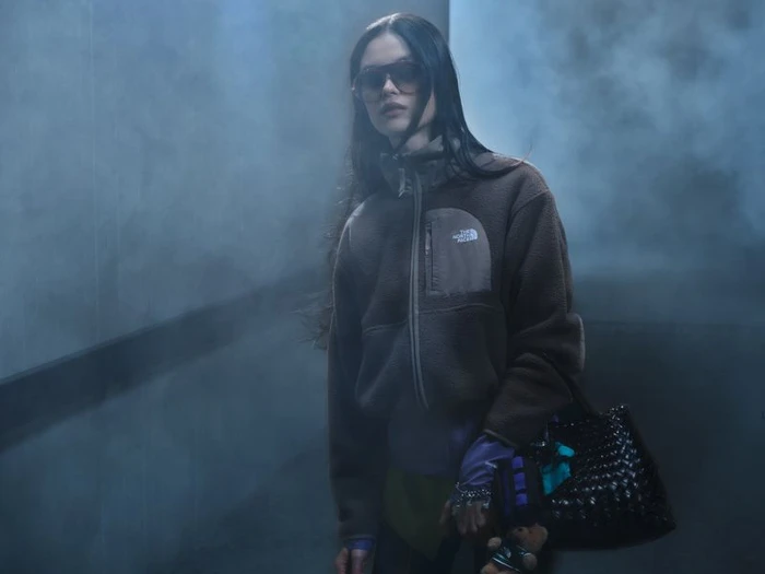 Model wearing dark sunglasses and a brown fleece jacket holding a black woven handbag amid a hazy foggy atmosphere.