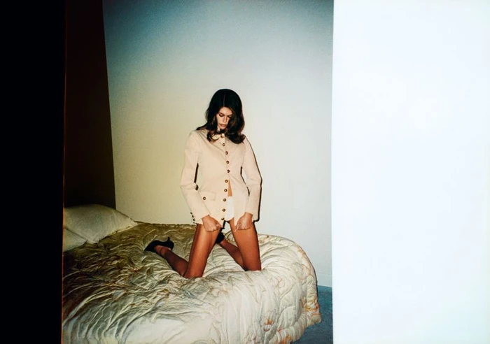 Model Kaia Gerber kneeling on a quilted bed in a buttoned jacket for RE/DONE