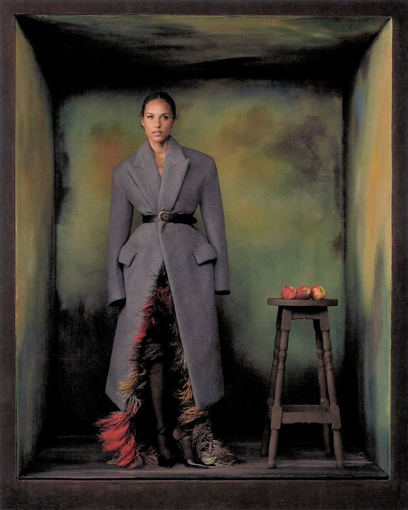 Alicia Keys standing inside a dark painted box wearing a grey coat and feathered skirt next to a wooden stool with apples