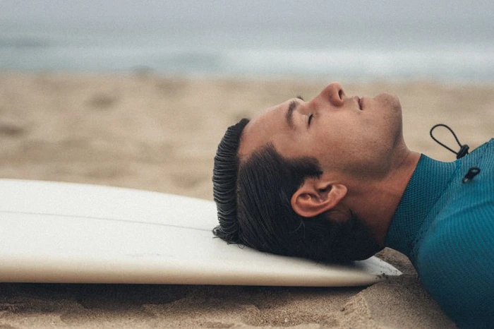Close-up profile of a man lying on a surfboard resting his head on it on a sandy beach